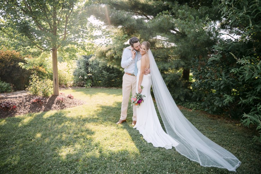 A groom kissing his bride’s cheek in a sunlit garden, her veil trailing across the grass